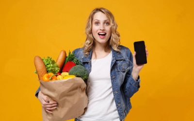 Excited girl in denim clothes isolated on orange background. Delivery service from shop or restaurant concept. Hold brown craft paper bag for takeaway with food product mobile phone with blank screen